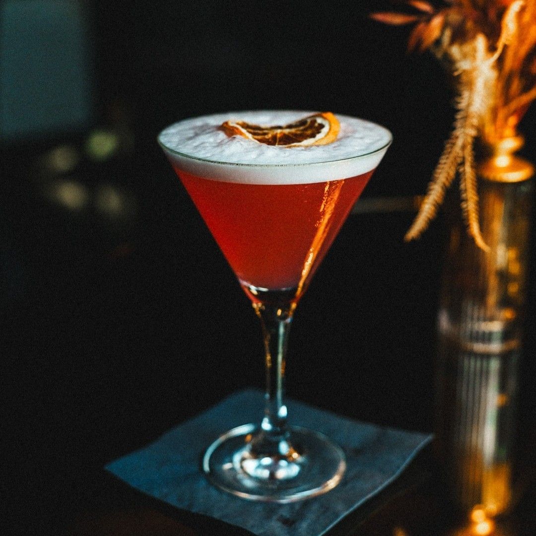 Red cocktail with white foam and citrus peel garnish in martini glass.