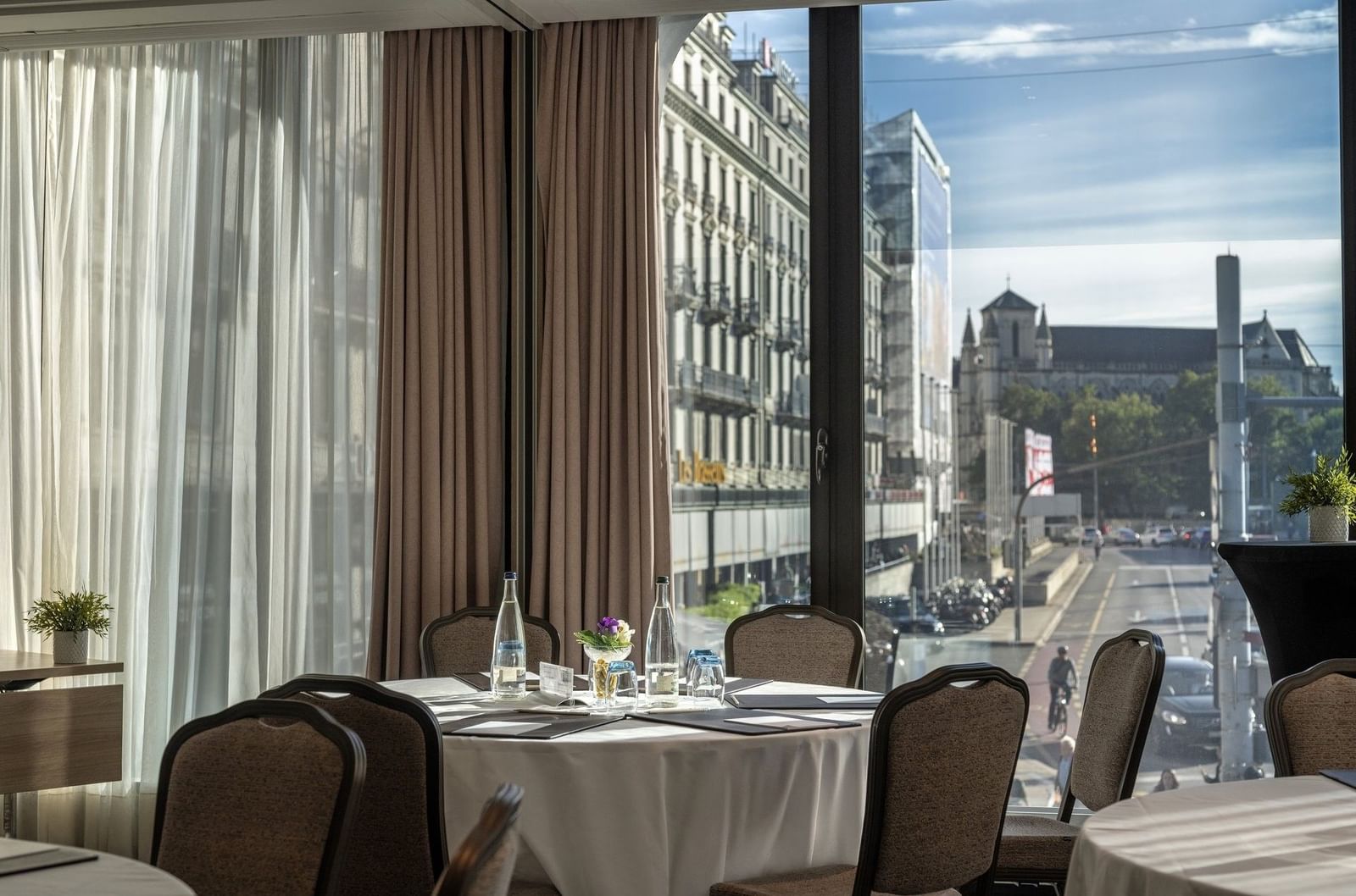 Round table with white linen by chairs under a curtained window near city views in Rigi + Cervin at Warwick Geneva