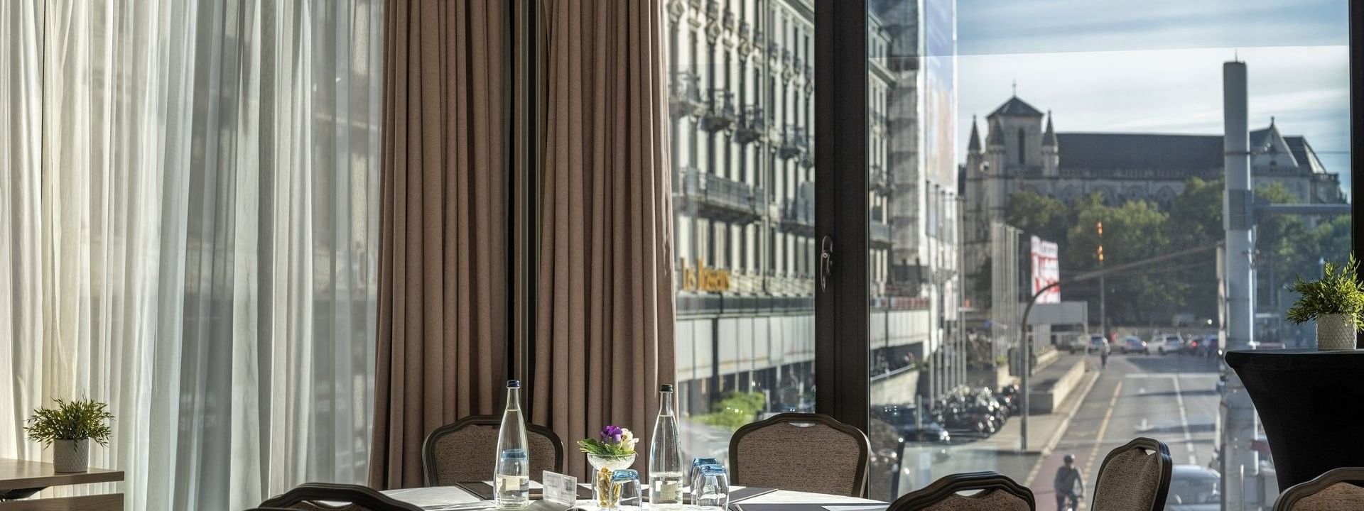 Round table with white linen by chairs under a curtained window near city views in Rigi + Cervin at Warwick Geneva