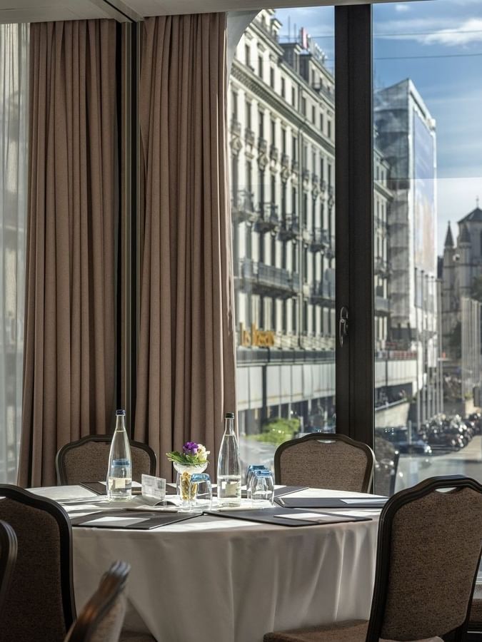 Round table with white linen by chairs under a curtained window near city views in Rigi + Cervin at Warwick Geneva