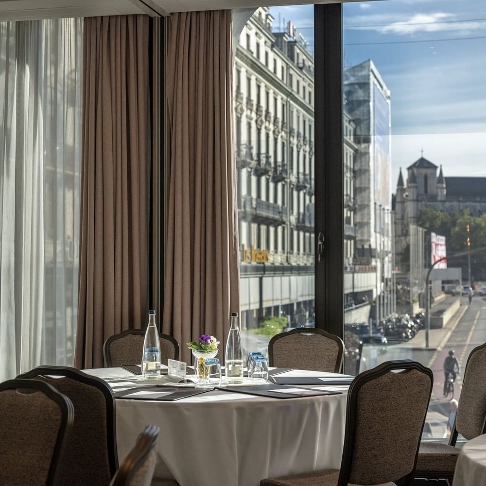 Round table with white linen by chairs under a curtained window near city views in Rigi + Cervin at Warwick Geneva