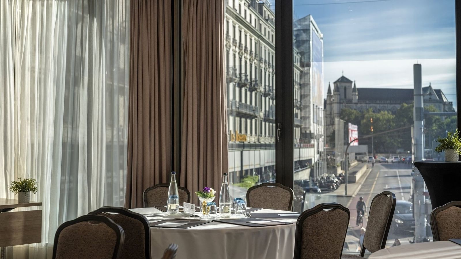 Round table with white linen by chairs under a curtained window near city views in Rigi + Cervin at Warwick Geneva
