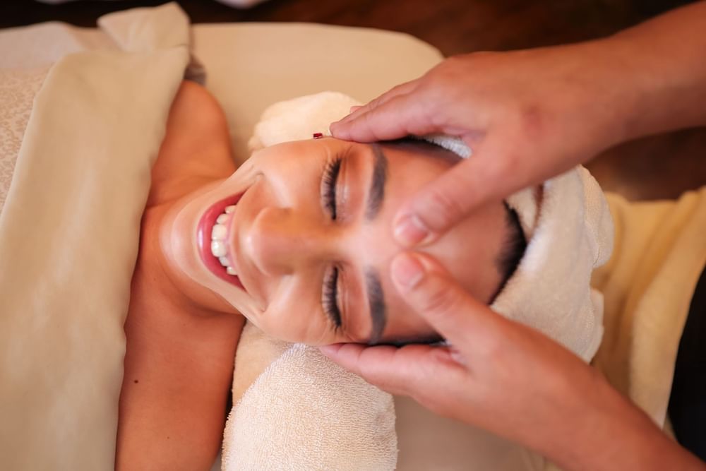 Close-up of a smiling woman receiving a relaxing forehead massage from a spa therapist's hands at Kelly's Spa