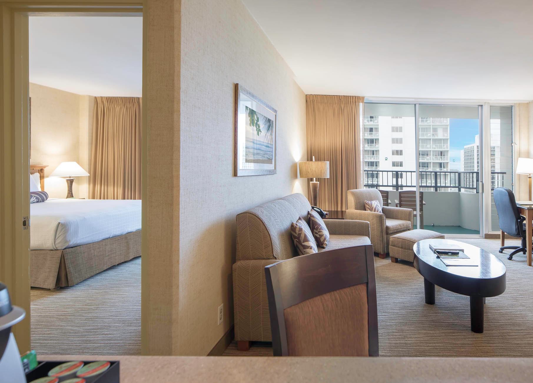A wide-angle view of a hotel suite with a living area, balcony access, and a bedroom in the background.