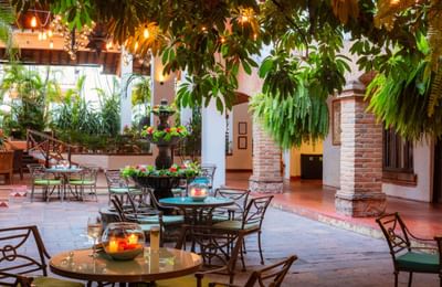 Outdoor patio with tables, chairs, brick columns, hanging plants, a fountain, and candles in the center.