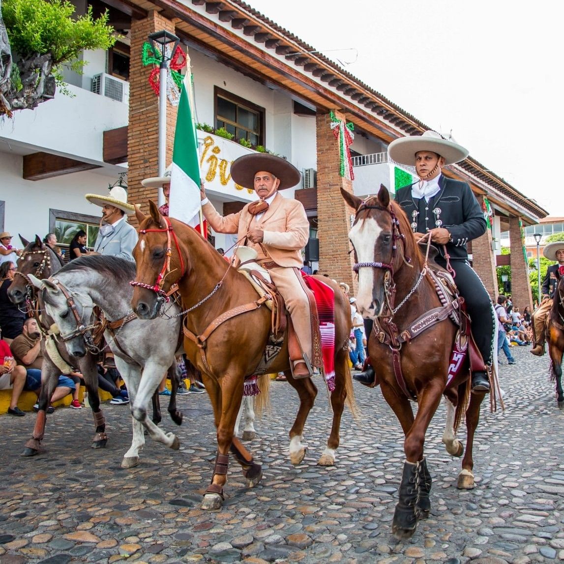 Charros en trajes típicos montando caballos en el día de la independencia de México callejera con espectadores y banderas.