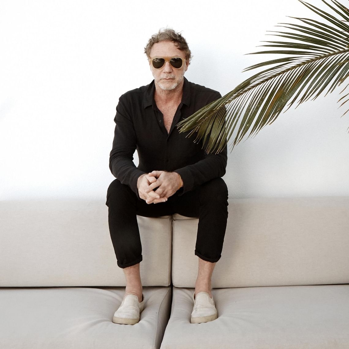 Oskar Metsavaht sitting on a couch wearing sunglasses with a palm frond in the foreground