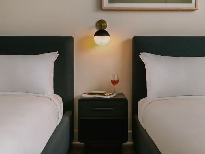 A nightstand with a glass of red wine between two luxurious beds and a modern lamp at Kenmore Village Hotel South Beach