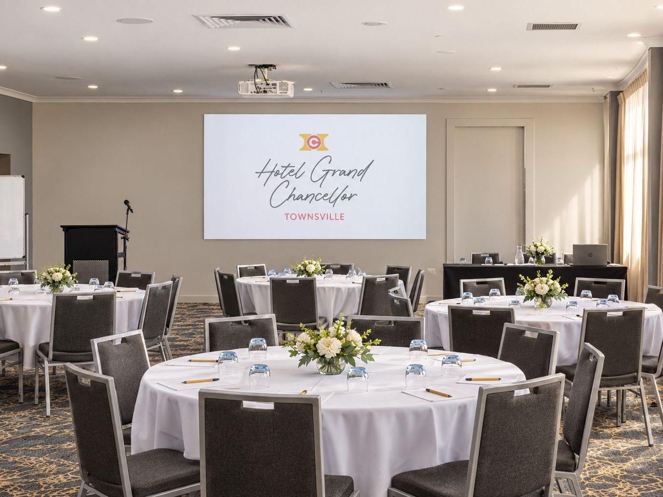 Conference room at Hotel Grand Chancellor Townsville featuring round tables with white linen, neatly arranged chairs, presentation facilities, projector screen and natural light from floor-to-ceiling windows.