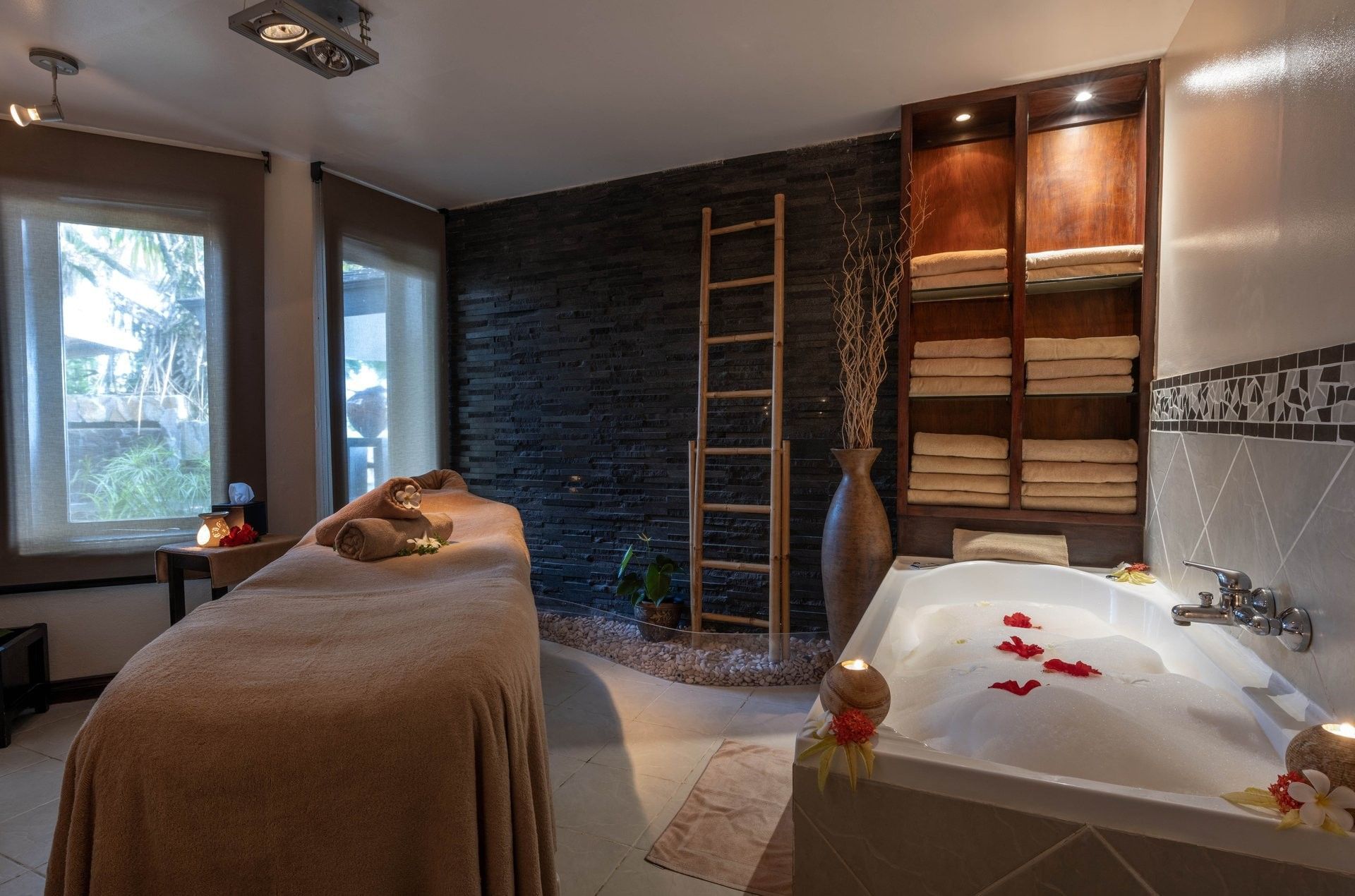Massage bed by a bathtub with petals under warm lights near a dark stone wall at Warwick Fiji Resort and Spa