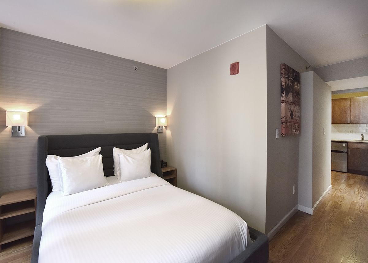 A hotel room with a bed, nightstands, lamps, and a kitchenette in the background.