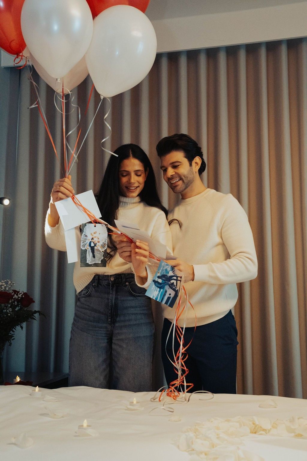 Couple reading photo notes tied to balloon ribbons, by a bed scattered with petals at Hotel X Toronto