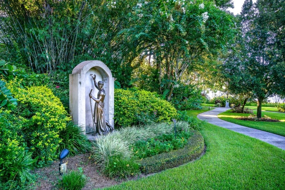 Lush gardens and trees surrounding a winding sidewalk with an arched stone feature with a bronze sculpture of a man in a robe with a walking stick.