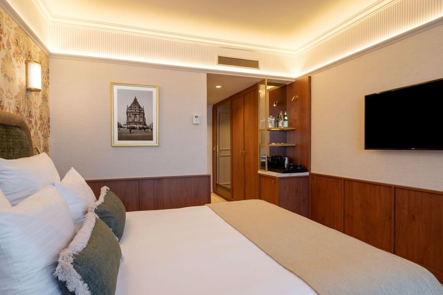 Modern hotel room with white bedding, wood paneling, minibar area, wall-mounted TV, and warm ambient lighting at Warwick Brussels Grand-Place