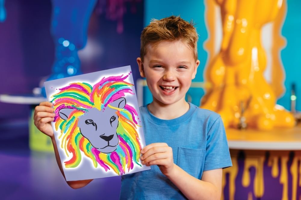 A smiling boy holds a colorful picture of a lion in a bright, colorful room. 