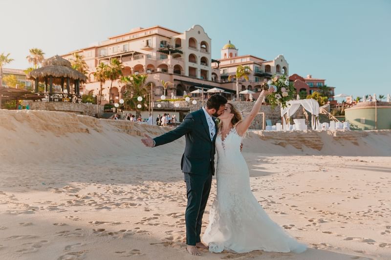 Pareja de recién casados besándose en la playa frente al edificio principal de Hacienda del Mar Los Cabos.