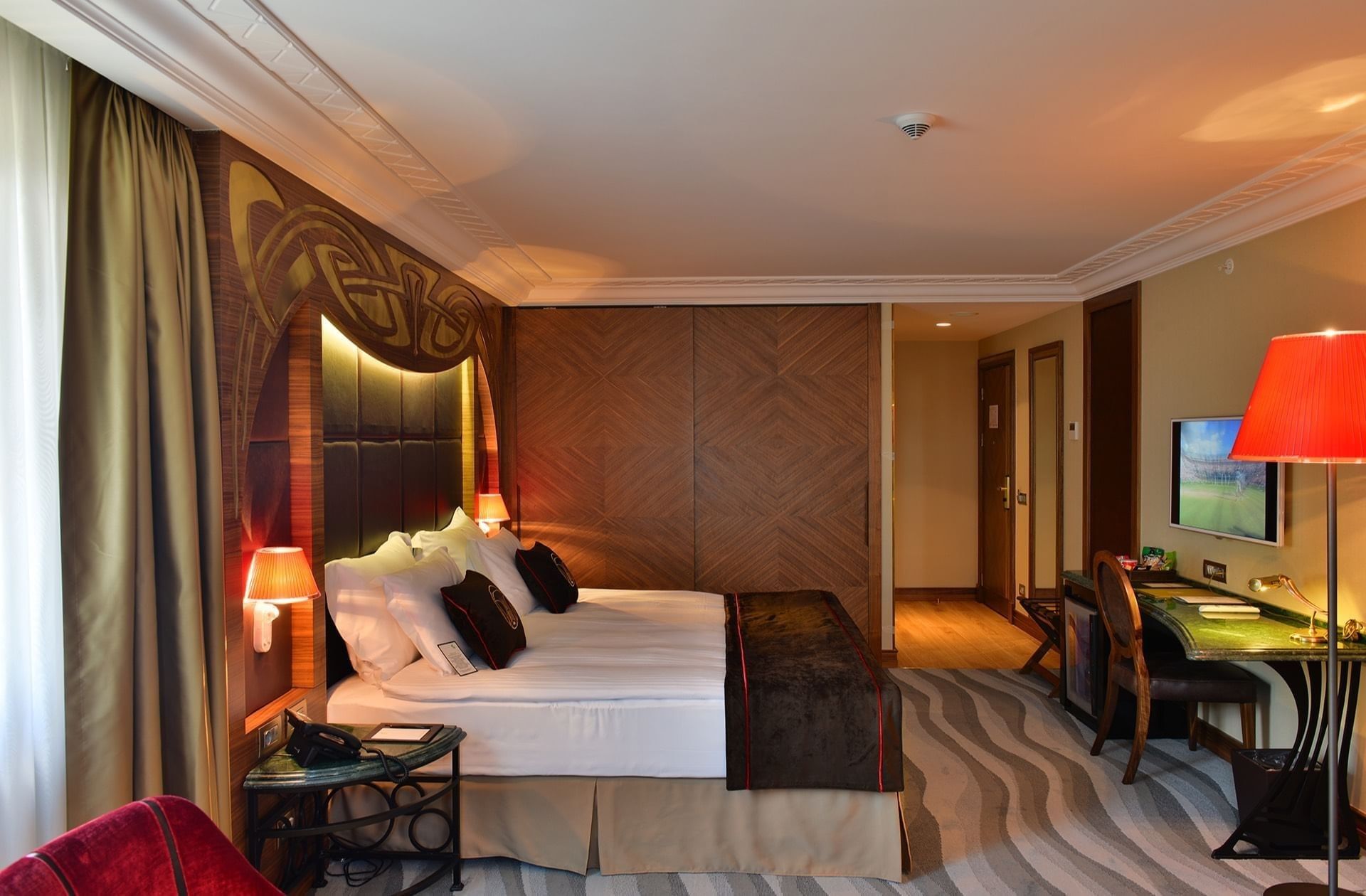 Work desk by a large bed with brown throws under a warm glowing light in Junior Suite at Warwick Ankara