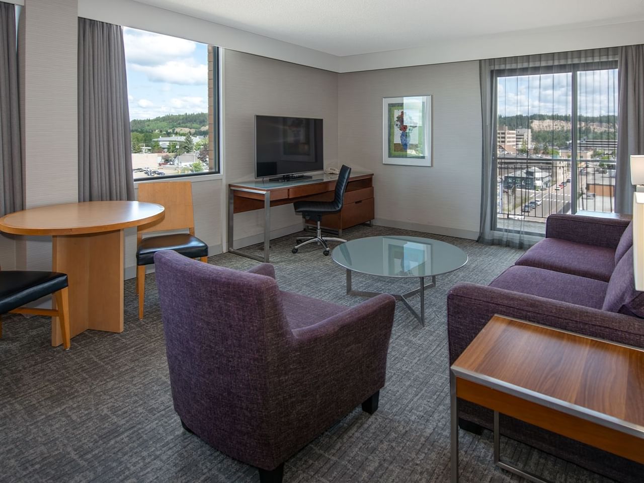 Living area with sofa, chair, desk, TV, and balcony in a hotel suite.