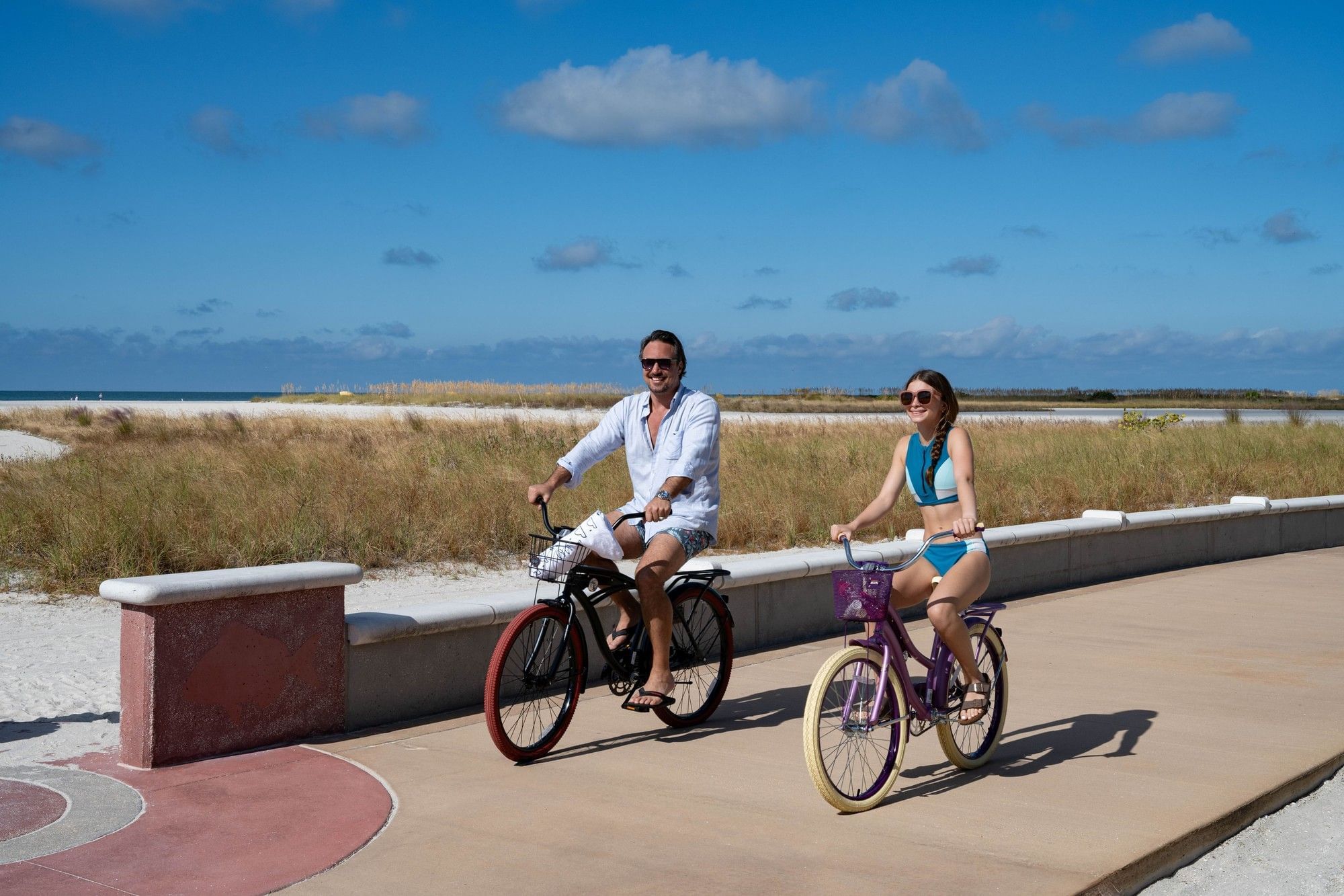 A couple rides bicycles along a beachside path with grass and a blue sky in the background.