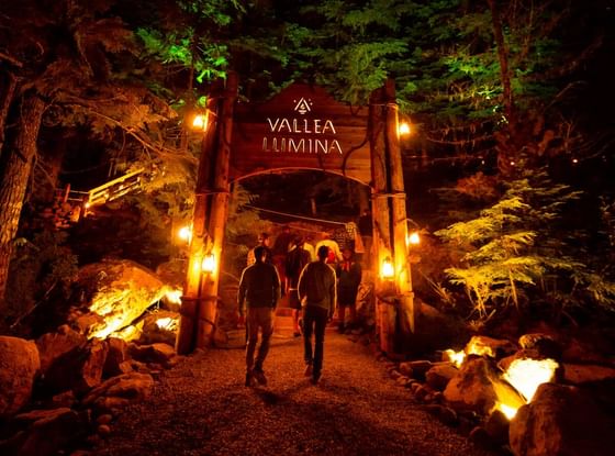 People walking under Vallea Lumina entrance arch at night surrounded by trees at Aava Whistler