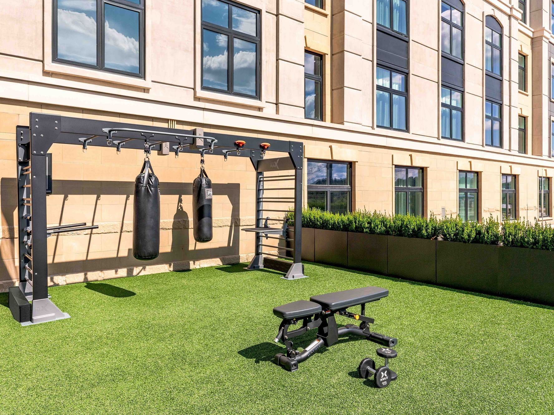 Outdoor gym setup with weight rack, punching bags, bench, and dumbbells on green turf against a building.