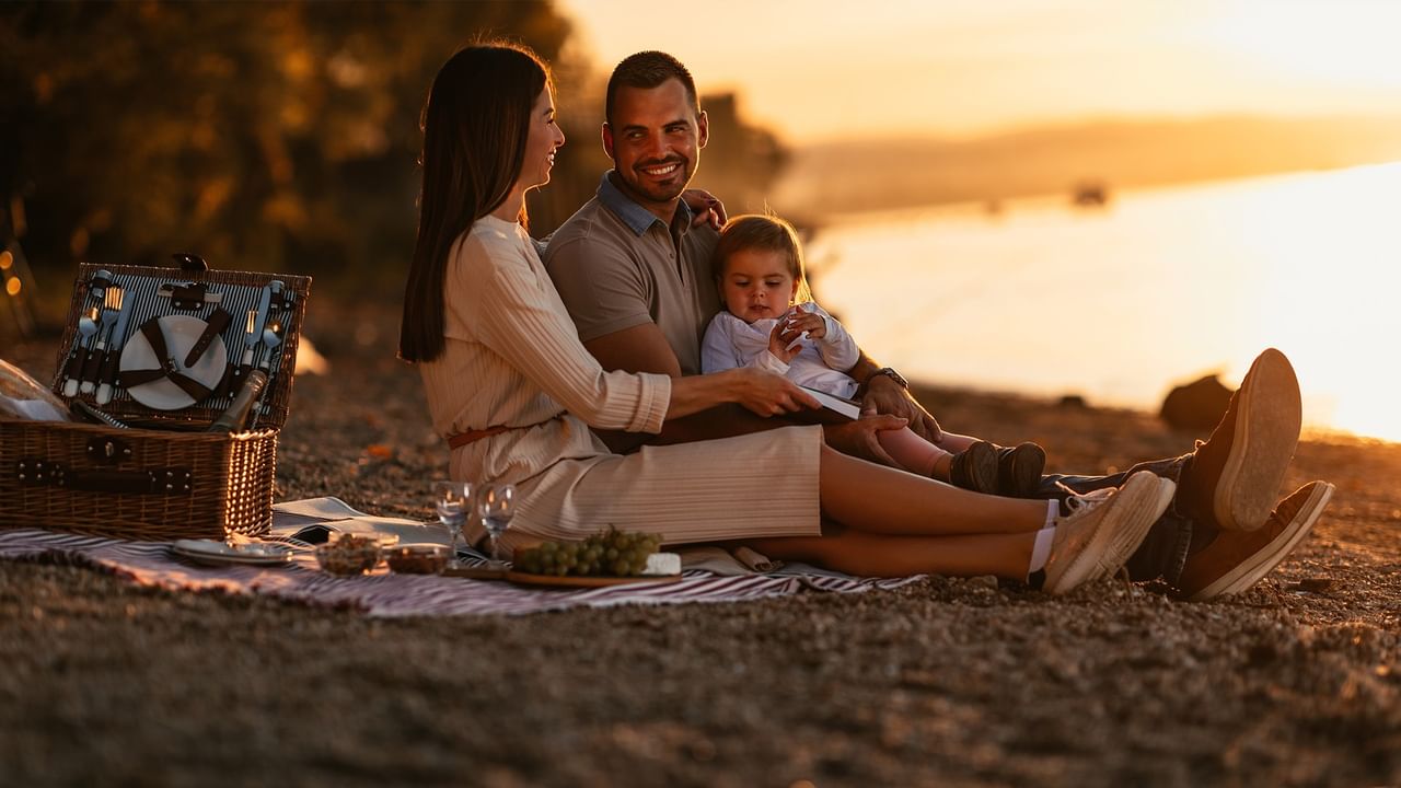 Family of three with picnic basket on beach at sunset, smiling and enjoying time together.