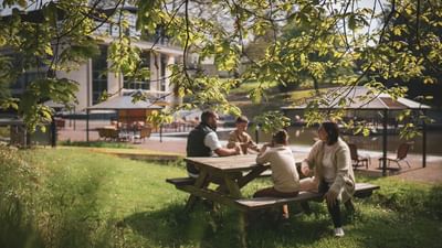 Four people sit at a picnic table in a park, with a lake and buildings in the background.
