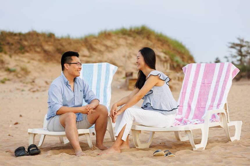 Couple on lounge chairs with striped towels talking on a sandy beach at Rodd Crowbush Golf & Beach Resort