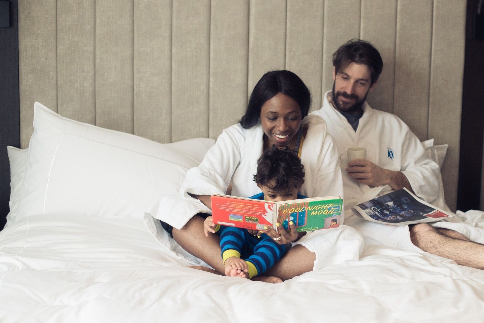 Family in white bathrobes sitting on plush king bed with young child reading storybook together at Hotel X Toronto