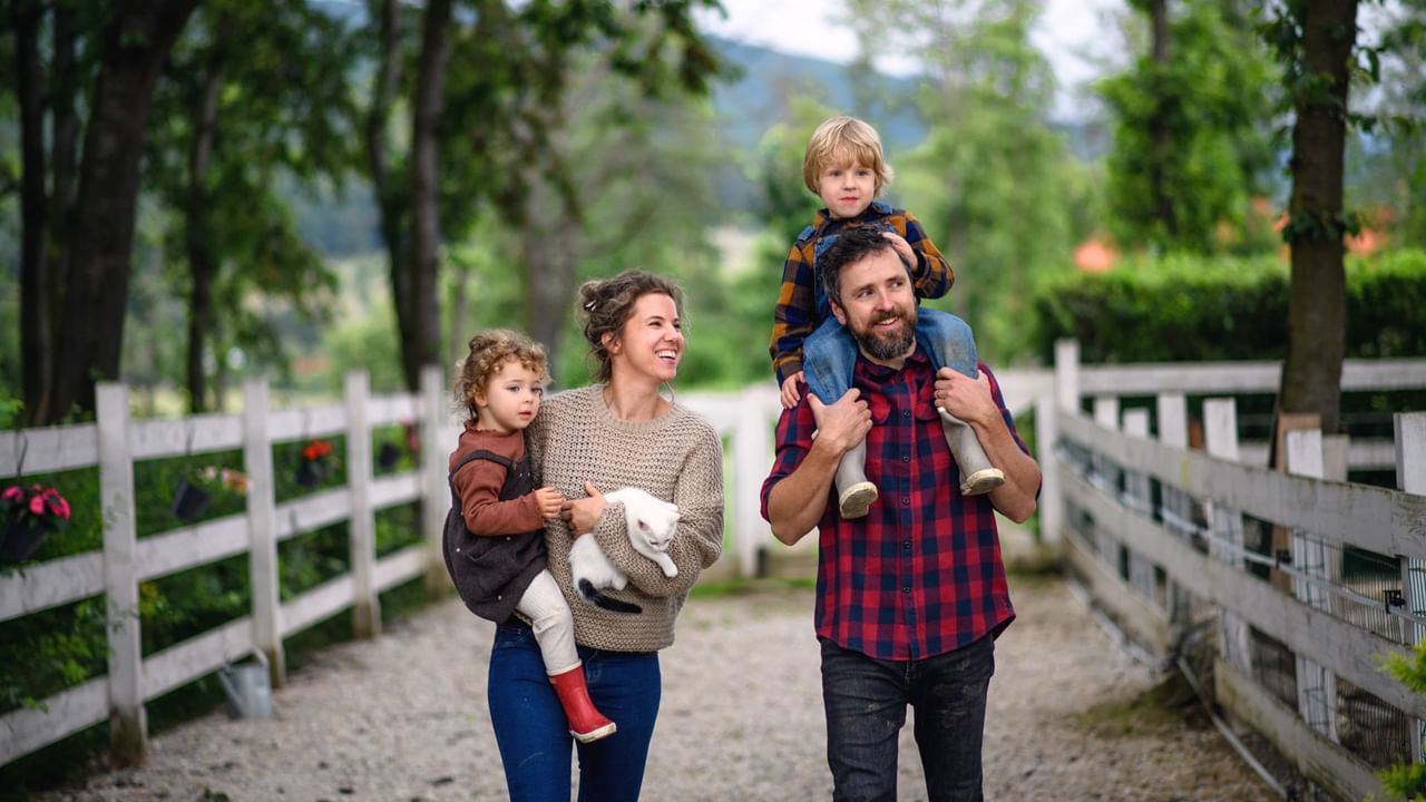 A smiling family enjoys a day at a farm, walking along a fenced path while holding a small white cat.