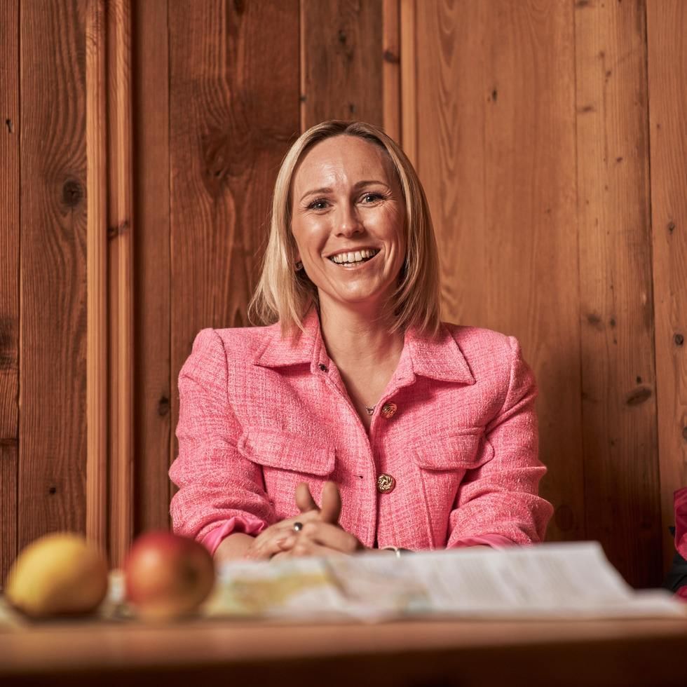 Eine lächelnde Frau in einem pinken Blazer sitzt an einem Tisch mit zwei Äpfeln.