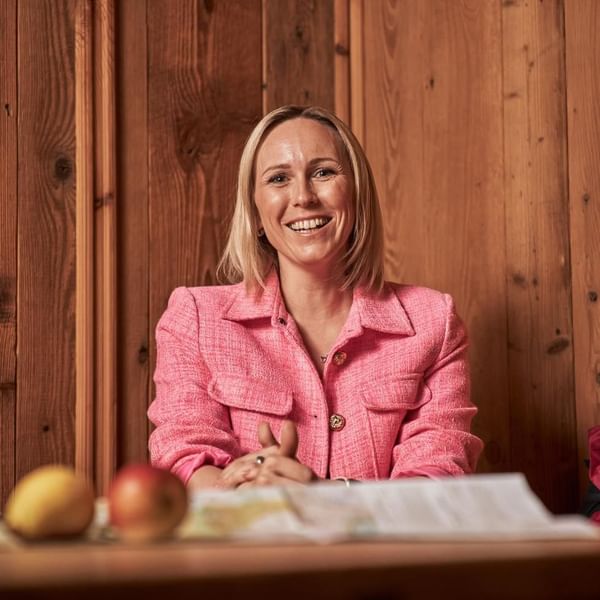 Eine lächelnde Frau in einem pinken Blazer sitzt an einem Tisch mit zwei Äpfeln.