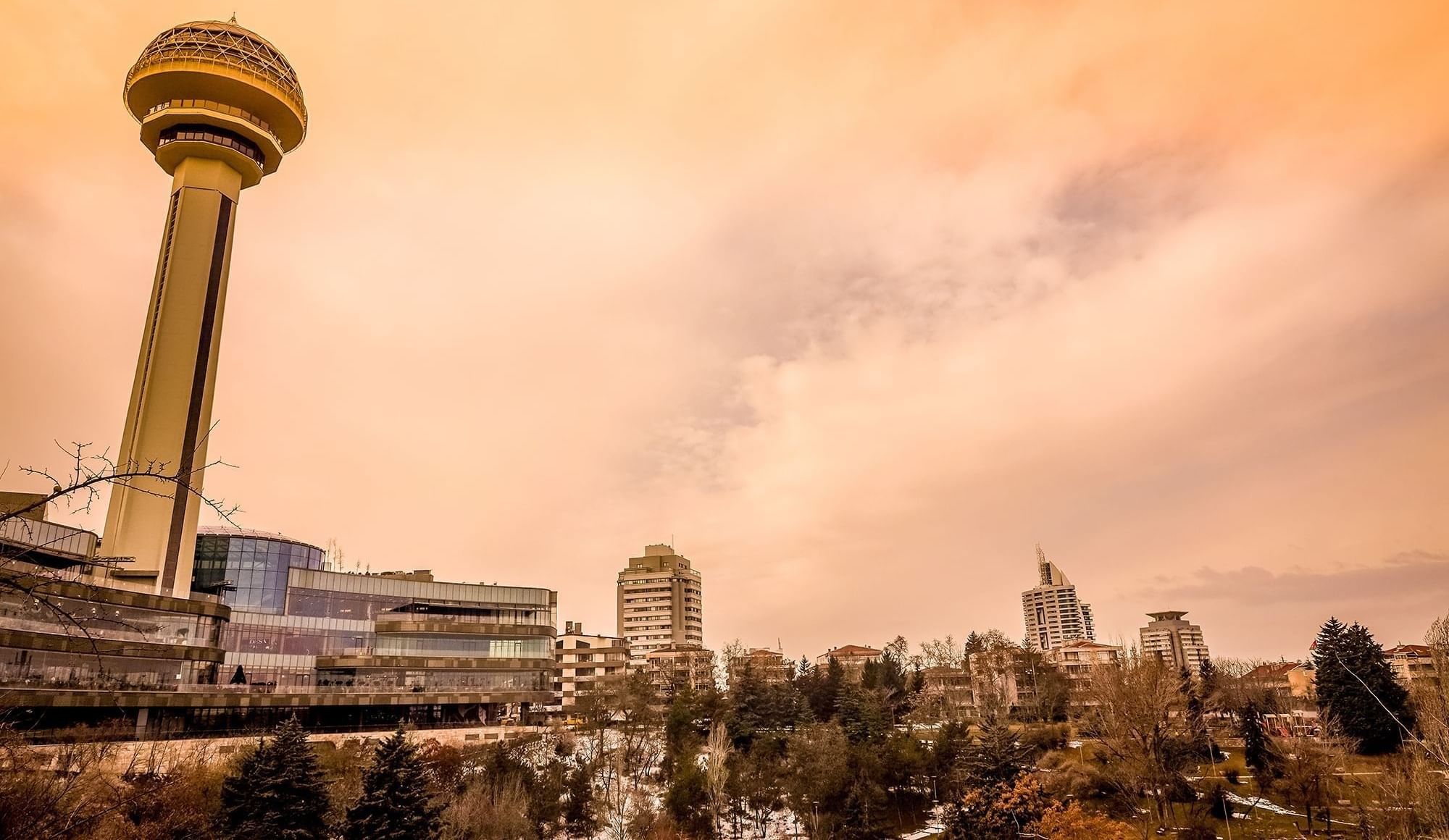 Atakule tower by a glass mall under an orange sunset sky near a wooded park area near Warwick Ankara