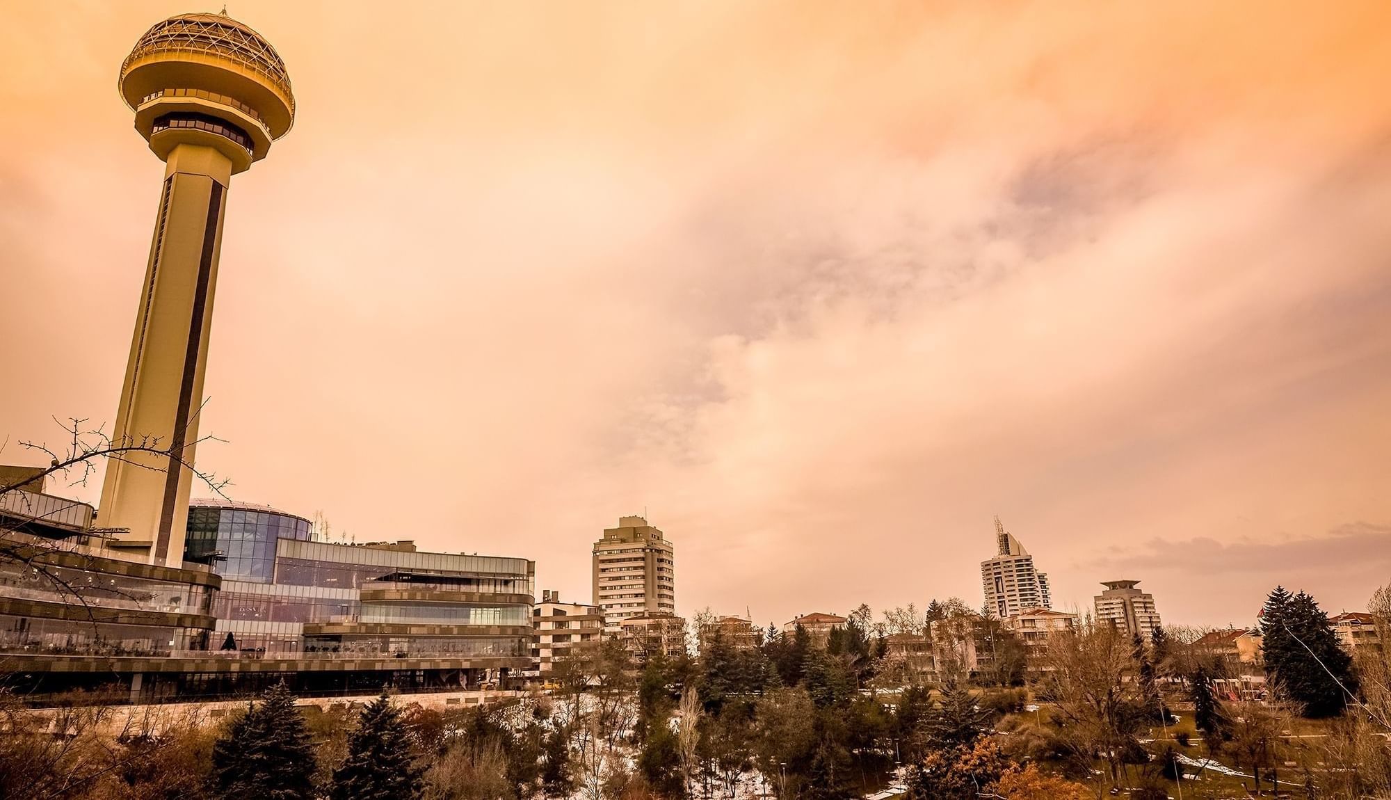 Atakule tower by a glass mall under an orange sunset sky near a wooded park area near Warwick Ankara