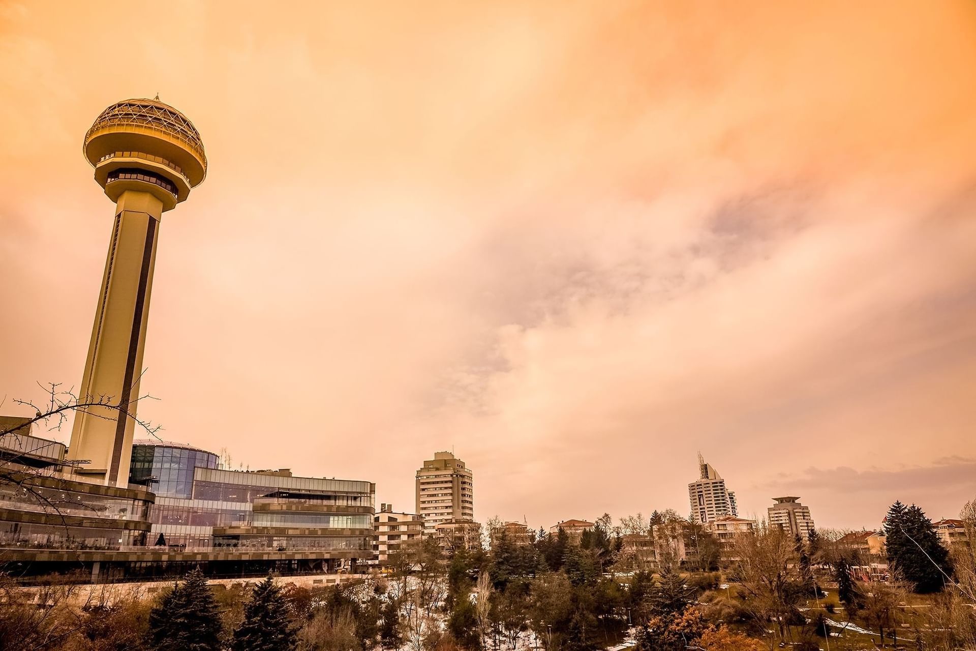 Atakule tower by a glass mall under an orange sunset sky near a wooded park area near Warwick Ankara