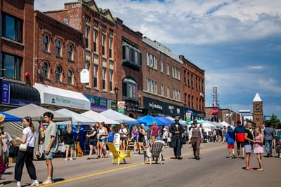 Crowd walking down a city street by vendor tents under a bright blue sky with clouds near Rodd Charlottetown Hotel