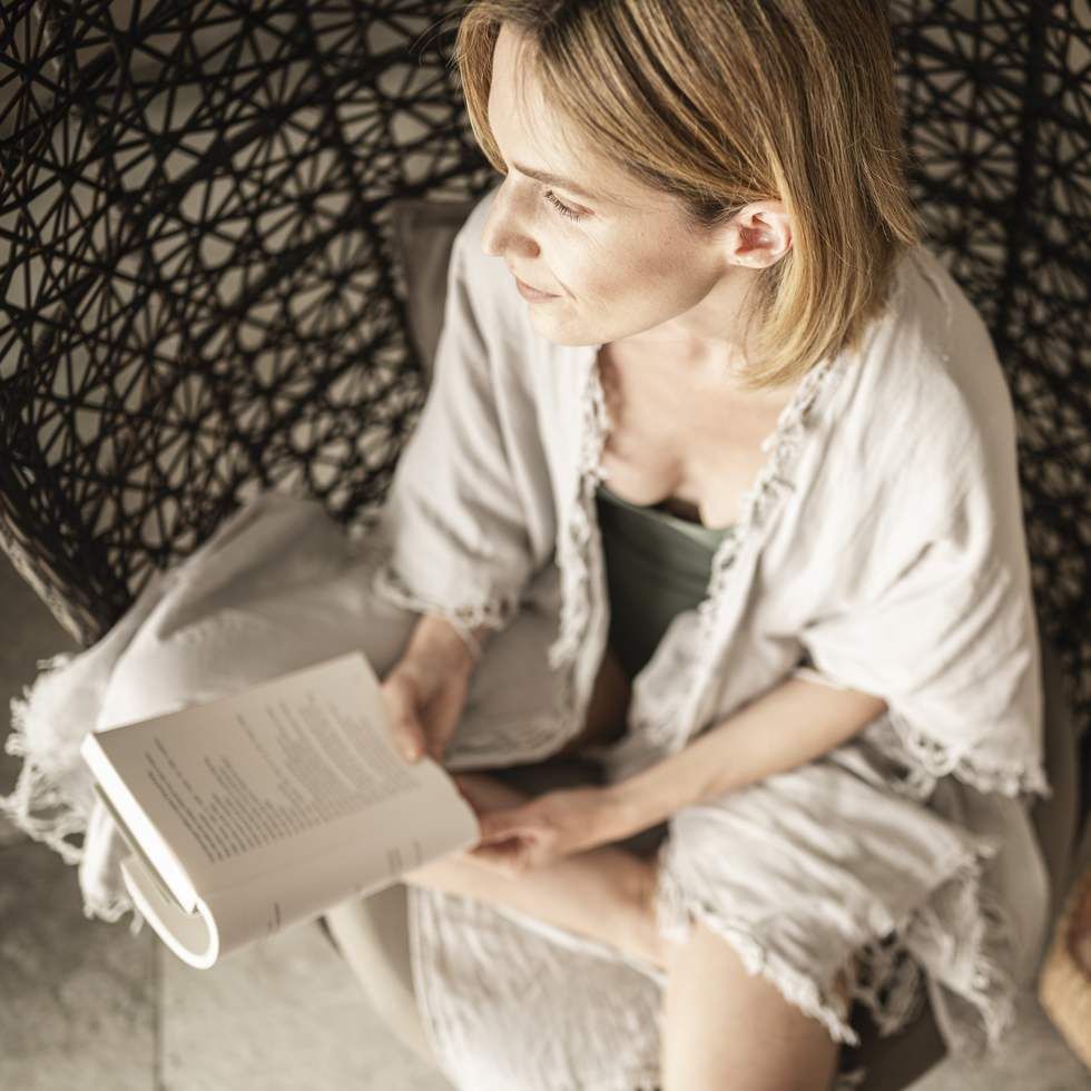 Frau mit blonden Haaren liest ein Buch in einem Korbsessel.