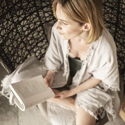Woman in a robe reading a book while sitting in a woven chair.