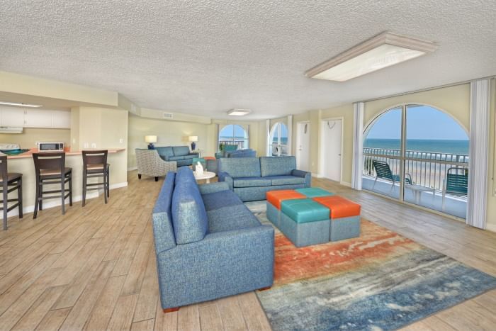 A wide view of a spacious living room with a kitchenette, two blue sofas, a coffee table, and a balcony with an ocean view.