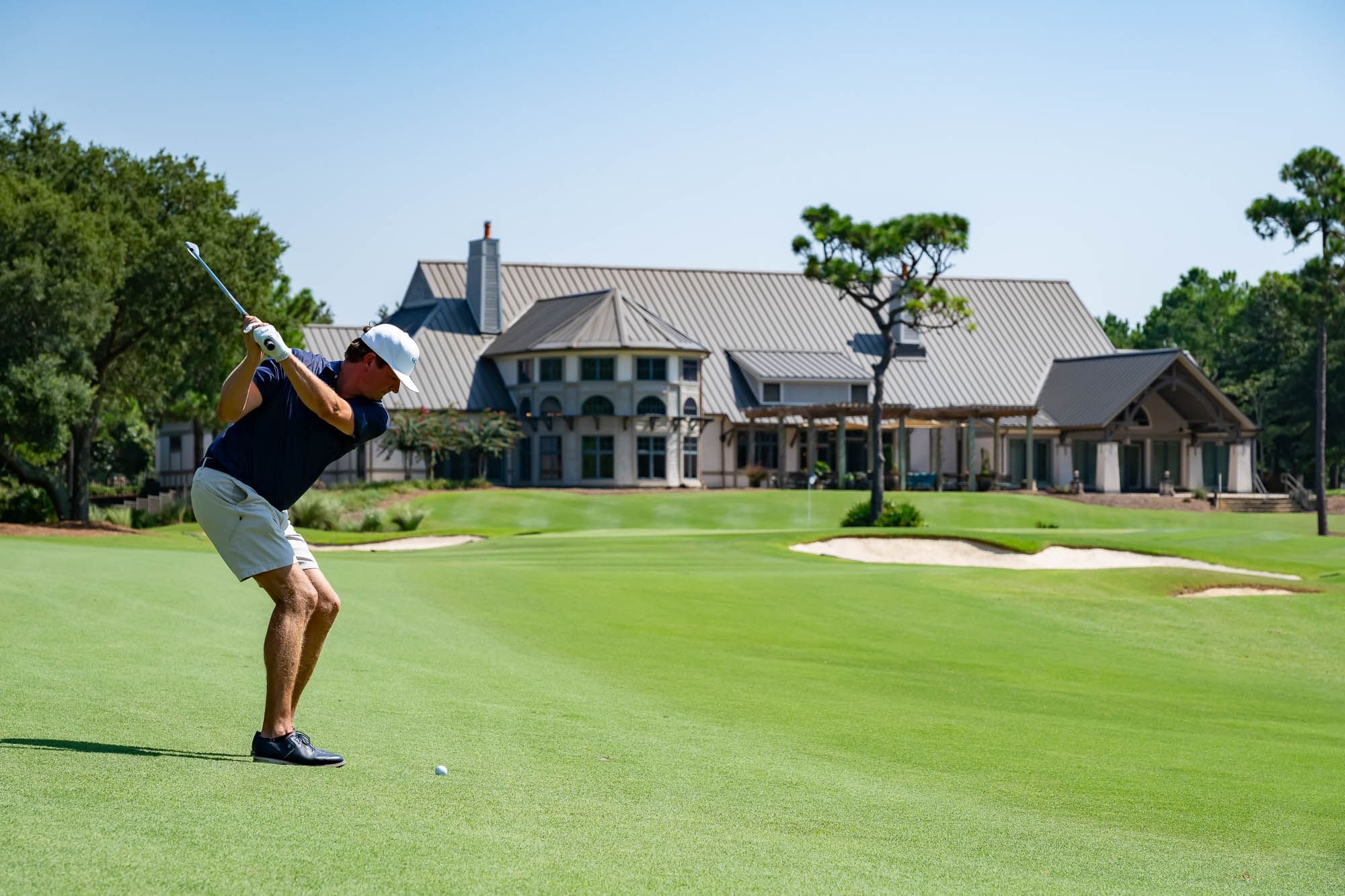 Golfer swinging club on a green course, with a large house and trees in the background.