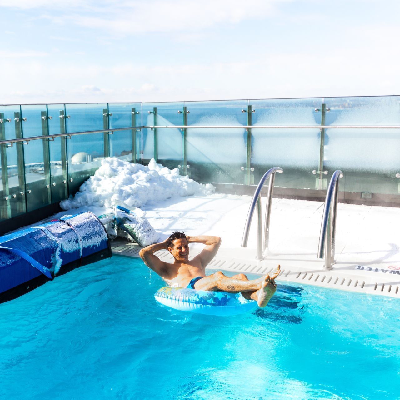 Man floating on an inflatable ring in a heated outdoor pool beside snow-covered decking at Hotel X Toronto