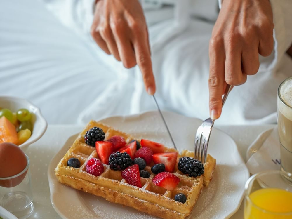 Guest cutting a Belgian waffle topped with fresh berries by a bowl of fruit at Warwick Grand Place Brussels