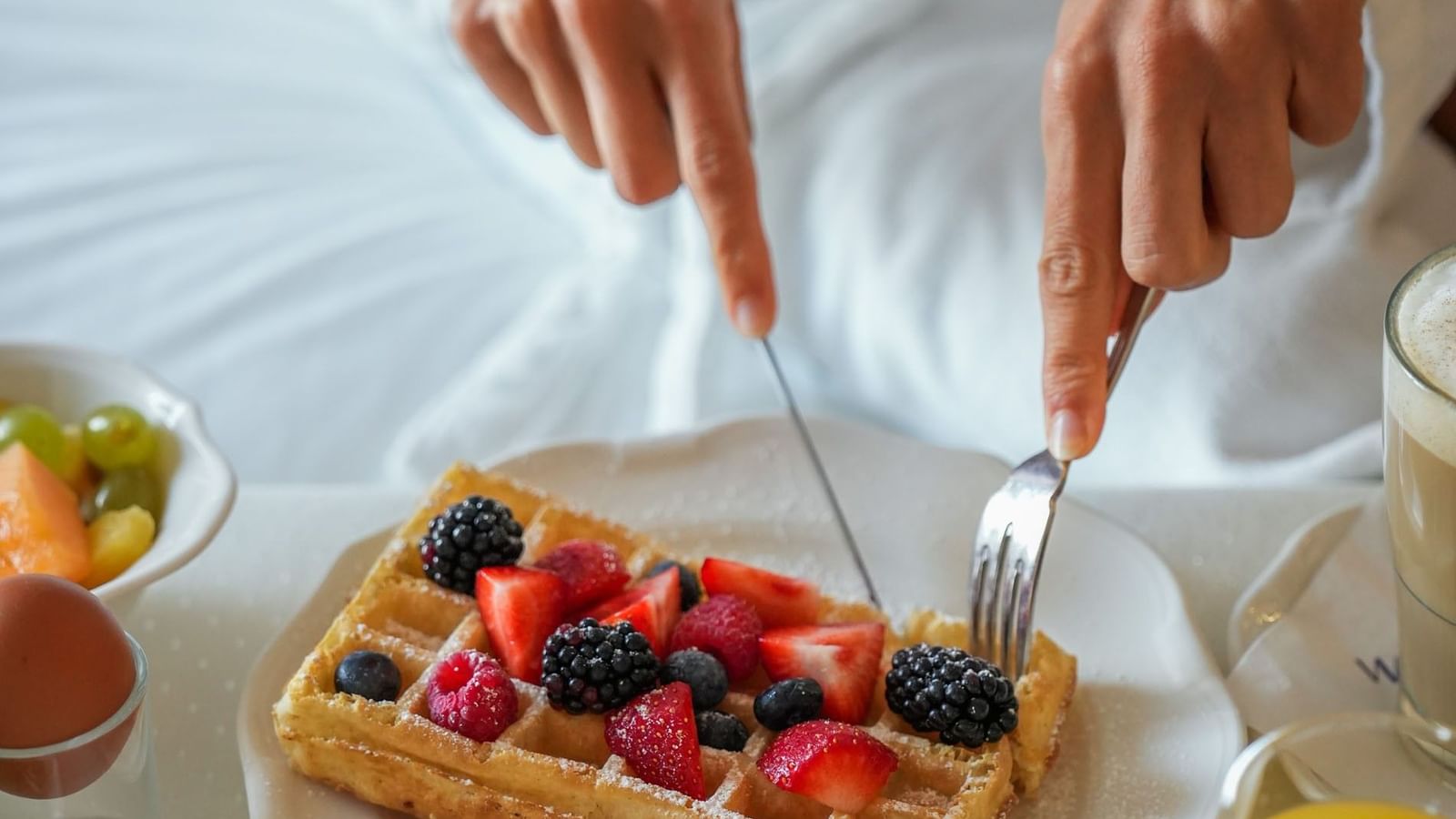 Guest cutting a Belgian waffle topped with fresh berries by a bowl of fruit at Warwick Grand Place Brussels
