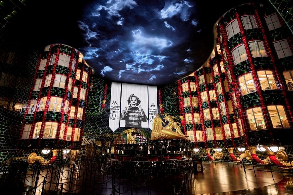 A dark lobby with a wall of towering windows surrounded by black brick, a large gold statue in the middle of the room, and the image of a woman screaming on a banner beneath stormy skies.