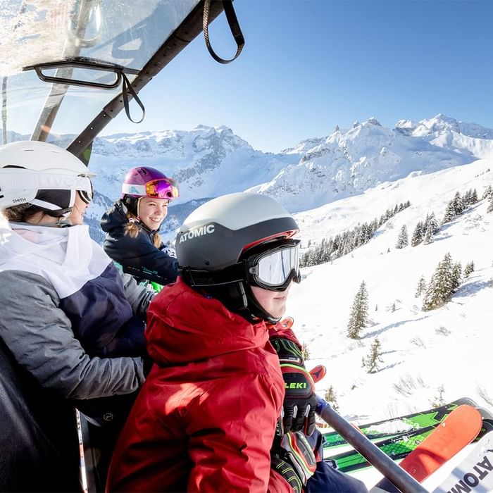 Drei Skifahrer in Skiausrüstung sitzen in einer Gondel mit Blick auf schneebedeckte Berge.
