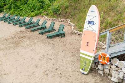 A paddleboard propped up on a sandy beach next to lounge chairs in The Lodge at Schroon Lake