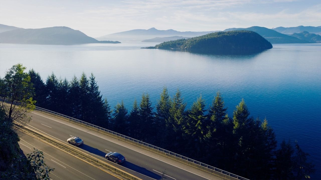 Two cars driving on a highway along a lake with mountains in the background.