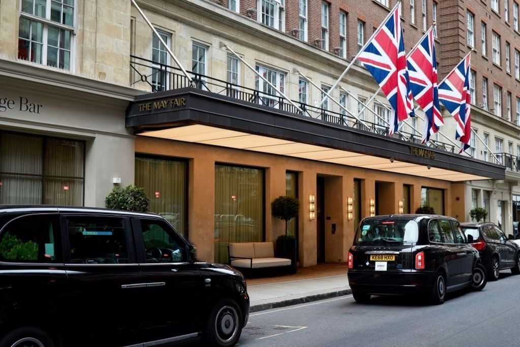 Front view of The May Fair Hotel entrance, adorned with Union Jack flags, and two black taxis parked outside