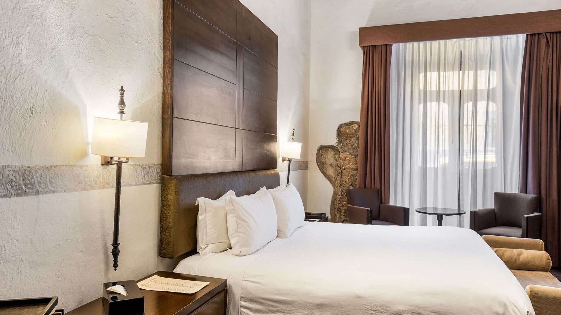 Luxury bed with a dark wood panel headboard and a decorative stone feature in the wall in King Superior at Quinta Real Puebla