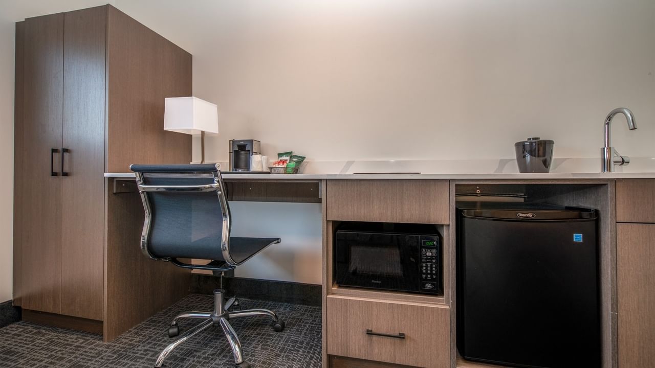 A hotel suite with a desk, chair, lamp, microwave, fridge, coffee machine, and a sink.